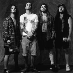 three men standing in front of a black and white background, one with long hair