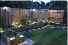 an outdoor garden area with wooden fence and raised planter boxes filled with plants, grass and lights