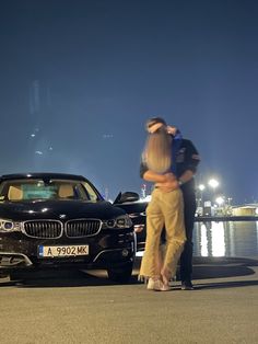 a man and woman standing next to a black car in front of a body of water