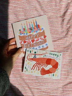 someone holding up two birthday cards on top of a bed with red and white striped sheets