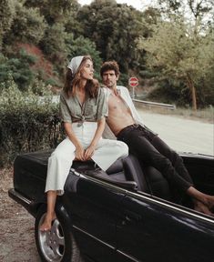 a man and woman sitting on the back of a black car in front of trees