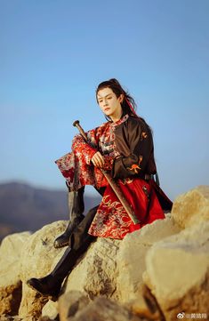 a woman sitting on top of a large rock with a stick in her hand and wearing a red dress