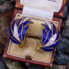 These ornate 18k yellow gold earrings feature a feathered design with blue enamel details. Three (3) of the feathers are accented with white rhodium plating and bead set with a total of nine (9) round single cut diamonds. The earrings measure 24.6mm long by 20.8mm wide and 13.1mm deep, and are finished with posts and omega backs for pierced ears. They are signed Muntejo.  One diamond is unnoticeably chipped. Luxury Hallmarked Enamel Earrings, Luxury Blue Enamel Earrings, Yellow Gold Earring, Feather Earrings, Rhodium Plated, Ear Piercings, Gold Earrings, Diamond Cuts, Yellow Gold