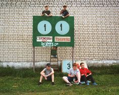 three boys are sitting on a sign in front of a brick wall and grass area