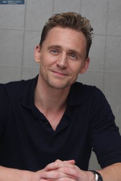 a man sitting at a table in front of a white wall with his hands folded out