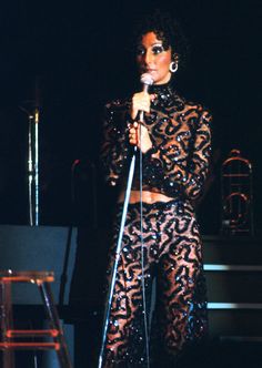 a woman standing in front of a microphone on stage wearing black and gold outfit with sequins