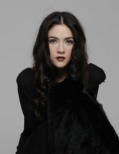 a woman with long dark hair wearing a black coat and posing for the camera in front of a gray background