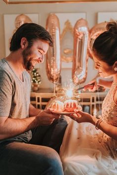 a man and woman sitting on a bed looking at a cake with candles in it