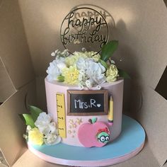 a birthday cake decorated with flowers, pencils and an apple is in a cardboard box