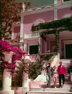 a man and woman standing on the steps in front of a pink house with stairs leading up to it