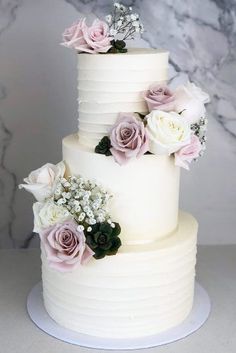 a three tiered white cake with pink and white flowers on top, sitting on a marble table