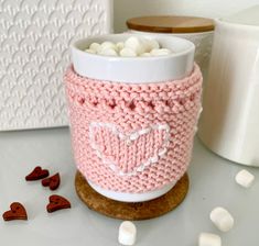 a knitted cup cozying up with marshmallows on the table next to it