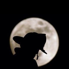 the silhouette of a man wearing a hat in front of a full moon