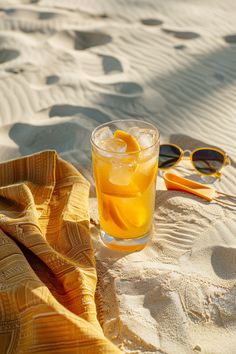 an orange drink sitting on top of a sandy beach next to sunglasses and a towel