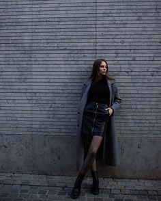 a woman leaning against a wall with her hand on her hip, wearing a gray coat and black boots