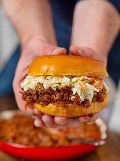 a person holding a sloppy joe sandwich over a bowl of chili and coleslaw