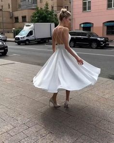 a woman in a white dress is standing on the sidewalk with her hands behind her back