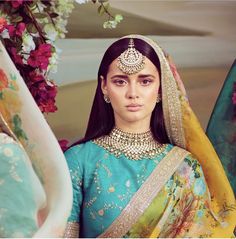 a woman in a green and yellow outfit with jewelry on her head standing next to flowers