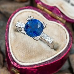 an oval blue sapphire and baguette diamond ring sits in a velvet box on top of rocks