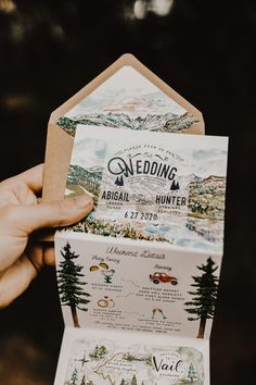 a person holding up a wedding card with mountains and trees on it in front of the camera