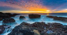 the sun is setting over the ocean with rocks in the foreground and waves crashing on the shore