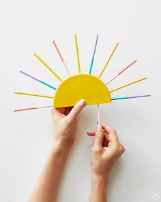 a person holding a yellow paper sun with colored sticks sticking out of it's center