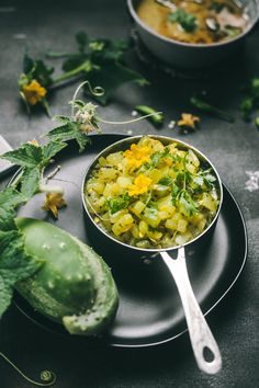 a black plate topped with a bowl of food next to cucumbers and flowers