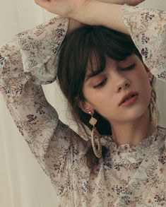 a woman with her hands behind her head wearing earrings and a floral blouse, standing in front of a white wall