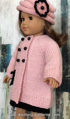 a doll wearing a pink knitted coat and hat with flowers on the side, standing next to a wooden wall