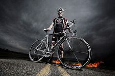 a man standing next to his bike on the road with flames in the air behind him