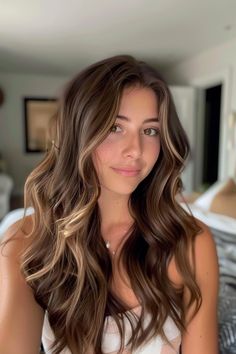 Young woman with long, wavy hair smiling in a cozy bedroom. Light Highlights In Dark Brown Hair, Golden Brown And Dark Brown Hair, Fall Brown Hair Highlights, Chocolate Lowlights On Blonde Hair, Brownie Hair With Caramel Highlights, Low Maintenance Brown Hair Balayage, Dimensional Brunette Level 6, Sunkissed Chocolate Brown Hair, Cute Brown Hair Highlights