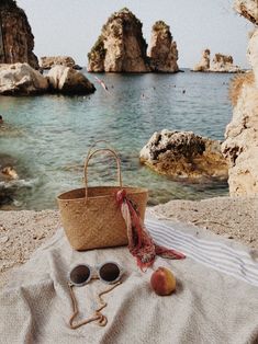 a basket and sunglasses on a towel next to the water with rocks in the background