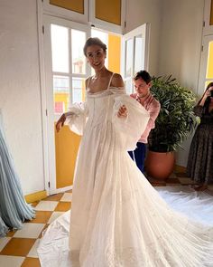 a woman in a white dress standing next to a window and another person taking pictures