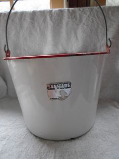 a white bucket sitting on top of a bed next to a window with a red handle