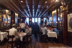 people sitting at tables in a restaurant with wood floors and exposed ceilings, eating food