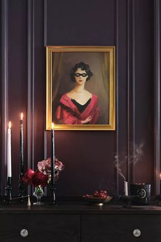 a portrait hangs on the wall above a dresser with candles and flowers in front of it