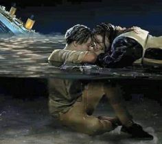 two people are kissing in the water next to a sinking boat and another person is sitting on the ground