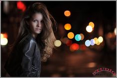 a woman with long hair standing in front of a street at night, wearing a black leather jacket