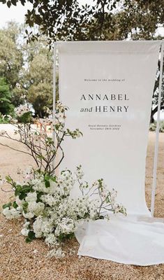 a white sign sitting on top of a sandy ground next to flowers and plants in front of it