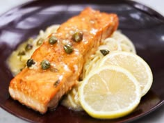 a piece of salmon with capers and lemons on a purple plate next to noodles