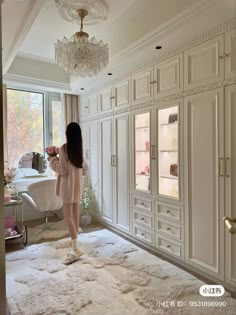 a woman standing in front of a large window next to a white cabinet and chandelier