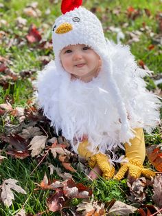 a baby wearing a chicken costume sitting in the grass with leaves all around her and smiling at the camera