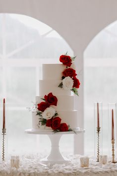 a white wedding cake with red and white flowers on top, surrounded by silver candles