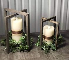 two white candles sitting next to each other on top of a wooden table with greenery