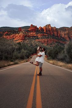 a man and woman walking down the middle of an empty road in front of mountains