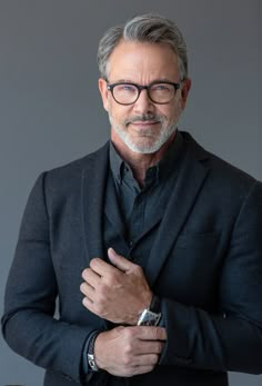 an older man wearing glasses and a black suit with his arms crossed, standing in front of a gray background