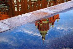 a reflection of a clock tower in the water on top of a sidewalk next to a building