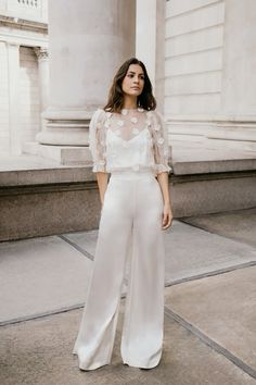 a woman standing in front of a building wearing white pants and a sheer top with flowers on it