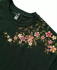 a green t - shirt with pink and yellow flowers embroidered on the chest, sitting on a white surface