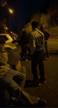 a group of people sitting on top of a sidewalk next to a white car at night